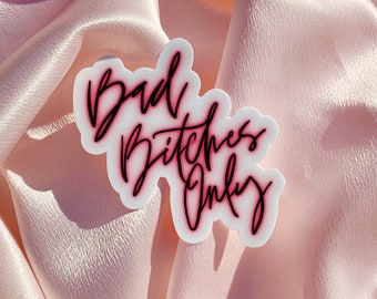 Può includere: Un adesivo bianco con la scritta "Bad Bitches Only" in corsivo nero con un contorno rosa. L'adesivo è posto su un tessuto di raso rosa chiaro.
