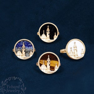 May include: Four gold rings with different colored stones. Each ring features a detailed white enamel design of a building with a tower.