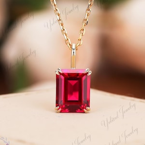 Könnte beinhalten: Eine goldfarbene Halskette mit einem rechteckigen, leuchtend roten Edelsteinanhänger. Der Anhänger ist in einer Vier-Krappen-Fassung gehalten und hängt an einer zarten Kette. Der Hintergrund ist unscharf.