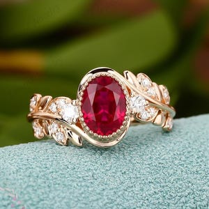 Peut inclure: Une bague en or avec une pierre rouge centrale de taille ovale entourée d'un bord perlé. L'anneau est orné de motifs de feuilles métalliques accentués par de petites pierres rondes transparentes incrustées le long de la tige.