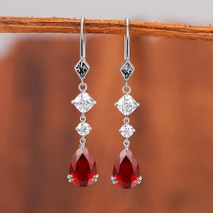May include: Silver drop earrings with a decorative black and silver top, a square white gem, a small round white gem, and a large teardrop-shaped red gem. The earrings are hanging from a brown branch.