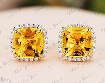 May include: A pair of square-cut, yellow gemstone stud earrings. Each earring features a large, vibrant yellow center stone surrounded by a halo of small, clear accent stones. The earrings are set in a gold-colored metal.