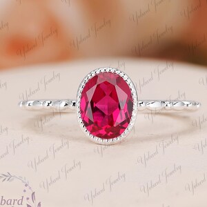 Puede incluir: Un anillo de plata con una gema ovalada de color rojo brillante. La gema está rodeada por un borde con cuentas. La banda tiene un diseño texturizado. El anillo se muestra sobre una superficie rosa pálido. El texto "Yvbard Jewelry".