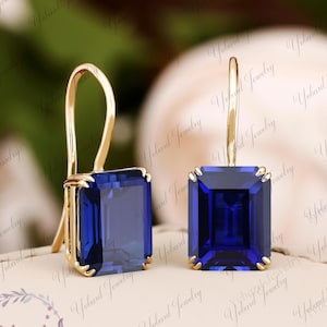 以下が含まれることがあります： 大きな長方形の濃い青色の宝石をあしらった、ゴールドカラーのドロップイヤリングのペア。イヤリングは、フック留めのシンプルなデザインです。宝石の長さは約1cmです。