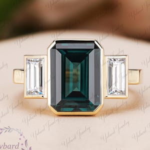 May include: A gold ring with a large emerald-cut teal gemstone and two rectangular white gemstones. The ring has a polished gold band. The jewellery is displayed on a light pink surface. The ring's design is simple and elegant.