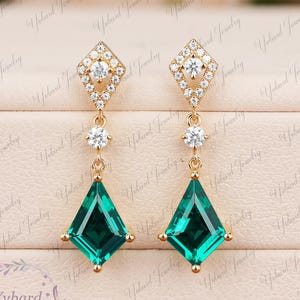 May include: A pair of gold drop earrings featuring a kite-shaped green gemstone suspended from a diamond-encrusted rhombus stud. Each earring includes a small round white stone connecting the stud to the larger green kite-cut stone.