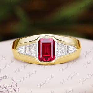 May include: A gold ring featuring a central emerald-cut red gemstone flanked by two trapezoid-cut clear stones on each side. The band is smooth and polished, showcasing a bezel setting that secures the vibrant red centre stone and the side accents.