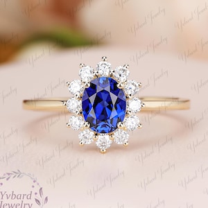 May include: A gold ring with a central, oval-cut, deep blue gemstone encircled by a halo of smaller, round, white gemstones. The ring has a slender gold band. The background is a soft pink hue.