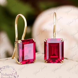 May include: A pair of gold-colored earrings with large, rectangular, ruby-red gemstones. The earrings have a hook design for pierced ears. The gemstones are set in a gold-colored metal. The earrings are displayed on a light-colored surface.
