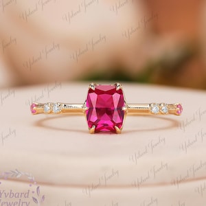 May include: A gold ring with a large, rectangular, bright pink gemstone as the centerpiece. The band is adorned with small diamonds and pink stones. The ring is set against a soft, neutral background. The text "Yvbard Jewelry" is visible.