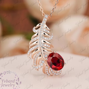 May include: A rose gold feather pendant necklace featuring a large, oval-cut, deep red gemstone. The feather design is adorned with small, sparkling clear stones. The necklace is displayed on a soft, pink surface.