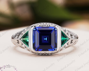 May include: A silver ring with a square blue gemstone and two triangular green gemstones on either side.