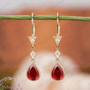 Puede incluir: Pendientes colgantes dorados con una piedra preciosa roja oscura en forma de lágrima. Los pendientes tienen un cierre de palanca y están adornados con pequeñas piedras de acento transparentes. El diseño incluye formas geométricas y una estética delicada.