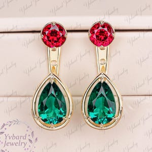 May include: A pair of gold-toned earrings featuring a round, faceted red stone at the top and a larger, pear-shaped, faceted green stone suspended within a gold-toned frame. The design includes a small circular cutout on the vertical connector.