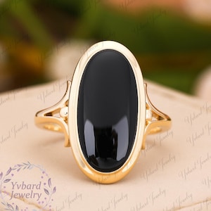 Könnte beinhalten: Ein goldener Ring mit einem großen ovalen schwarzen Onyx-Edelstein. Der Ring hat ein zartes, verziertes Design.