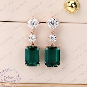 May include: A pair of dangle earrings with emerald green rectangular stones and clear round stones. The earrings are made of rose gold.