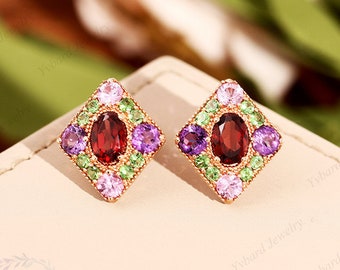 Könnte beinhalten: Ein Paar goldene Ohrringe mit quadratischem Design. Jeder Ohrring hat einen roten ovalen Edelstein, der von kleineren grünen, violetten und rosa Edelsteinen umgeben ist.