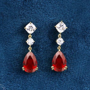 May include: A pair of gold-toned drop earrings. Each earring features a square white gemstone at the top, followed by a round white gemstone, and a large, teardrop-shaped red gemstone at the bottom. The earrings are set against a dark blue background.