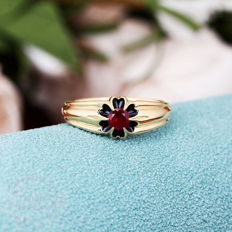 May include: A gold ring with a black flower design and a red gemstone in the center.