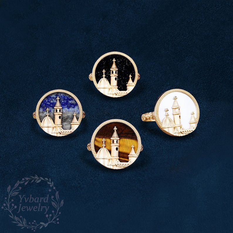May include: Four gold rings with different colored stones. Each ring features a detailed white enamel design of a building with a tower.