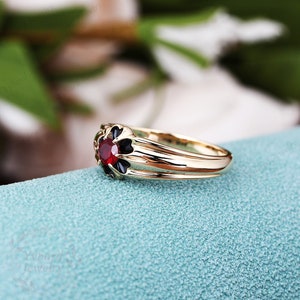 May include: A gold ring with a red gemstone surrounded by black enamel. The ring has a simple band with a slightly raised design.