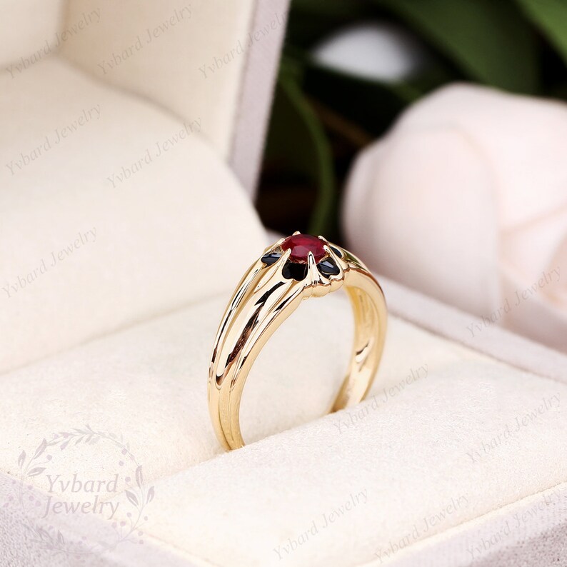 May include: A gold ring with a red gemstone surrounded by smaller black gemstones. The ring is in a white box.