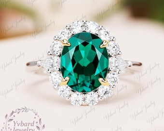 May include: A silver ring with a large oval emerald green gemstone surrounded by a halo of smaller round white gemstones. The ring has a delicate band with two smaller pear-shaped white gemstones on either side of the main stone.