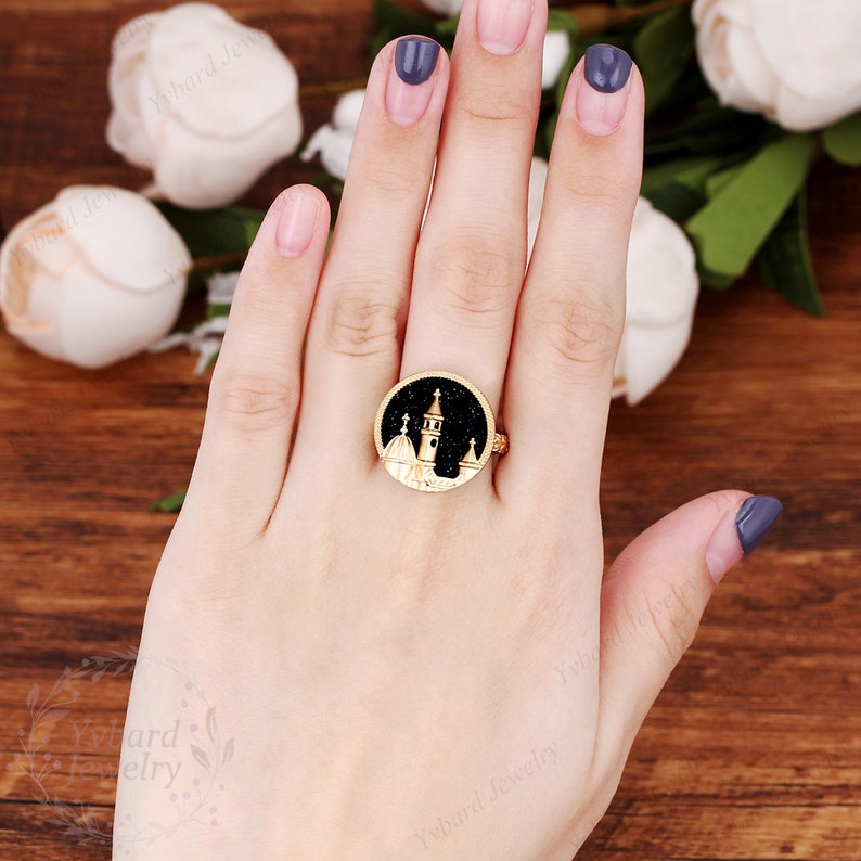 May include: Gold ring with a black enamel design of a building with white accents. The ring is on a person's finger.