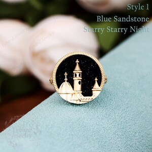 May include: A gold ring with a blue sandstone inlay depicting a cityscape at night. The ring features a detailed design of buildings with domes and a tall tower, all silhouetted against a starry sky.