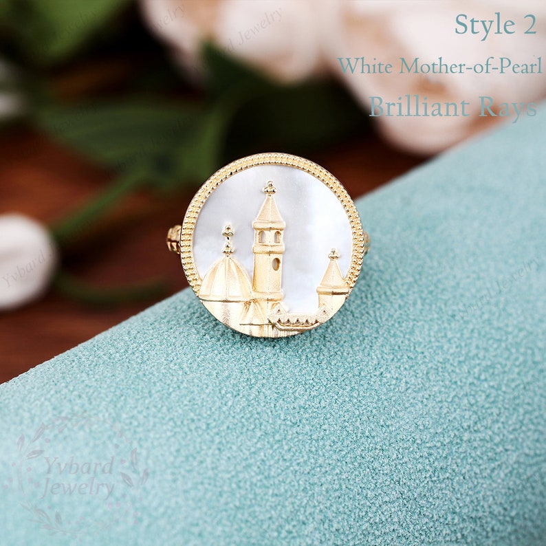 May include: A gold ring with a white mother-of-pearl inlay featuring a detailed design of a church with a tower and domes. The ring is on a light blue background.