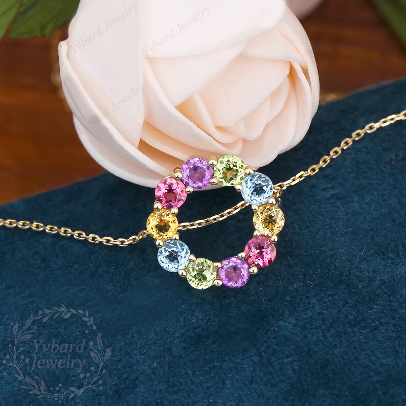 May include: A gold chain necklace with a colorful gemstone circle pendant. The pendant features a circle of round, faceted gemstones in shades of pink, purple, blue, green, and yellow.
