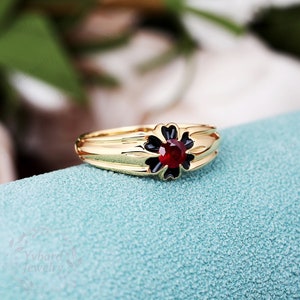 May include: A gold ring with a black flower design and a red gemstone in the center.