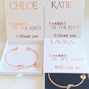 May include: Rose gold bracelets with a knot design and a small round charm, displayed in white boxes. The boxes feature rose gold foil text reading "CHLOE", "KATIE", and "LAURA", and the phrase "I couldn't tie the knot without you."