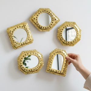 Puede incluir: Colección de cinco pequeños espejos decorativos dorados en varias formas geométricas, incluyendo un cuadrado, un hexágono y un óvalo. Cada espejo tiene un borde detallado y punteado y está montado en una pared blanca.