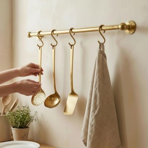 May include: A kitchen setting shows a brass-coloured utensil rack with hanging cooking tools. A slotted spoon, ladle, and spatula hang from S-hooks. A beige towel hangs on the right. White plates and a small potted plant sit on a wooden countertop.