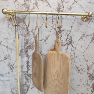 May include: A brass kitchen rail with hooks holding wooden cutting boards and a long, thin wooden utensil. The cutting boards are light-colored wood with natural grain patterns. The rail is mounted on a marble-patterned wall.