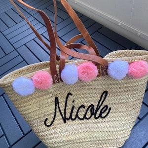 Puede incluir: Un bolso de paja con asas de cuero y pompones rosas y blancos alrededor de la parte superior. El bolso est&aacute; personalizado con el nombre "Nicole" en bordado negro.