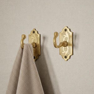 May include: Two antique brass wall hooks with a floral design. One hook holds a beige towel. The hooks are mounted on a light grey wall. The hooks are ornate and have a curved design.