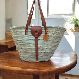 May include: A light blue woven straw tote bag with brown leather handles and a leather strap. The bag has a brown leather tag with the text "Art-Of-Handmade" on it.