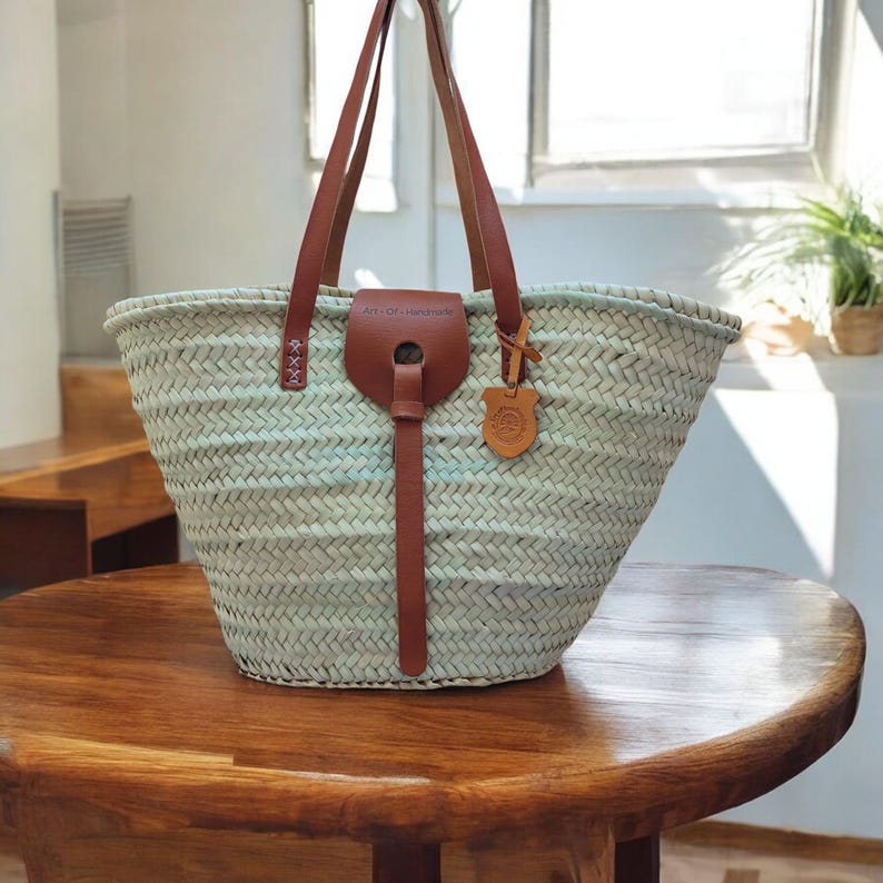 May include: A light blue woven straw tote bag with brown leather handles and a leather strap. The bag has a brown leather tag with the text "Art-Of-Handmade" on it.