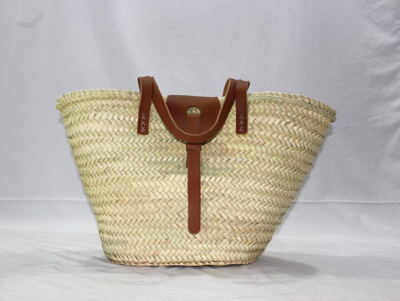 K&ouml;nnte beinhalten: Eine gro&szlig;e, geflochtene Strohtasche mit braunen Ledergriffen und einem braunen Lederriemen. Die Tasche ist aus nat&uuml;rlichem Stroh gefertigt und hat eine breite, offene Oberseite.
