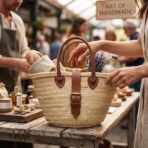 May include: A woven straw market basket featuring two brown leather handles and a central brown leather strap with a metal buckle. The basket contains a loaf of bread in a linen cloth, a bunch of dried lavender, and a small ceramic jug.
