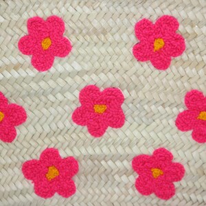 May include: Woven straw bag with bright pink flower appliqu&eacute;s. Each flower has a yellow center. The bag's weave is visible, creating a textured surface. The flowers are evenly spaced across the bag's surface.