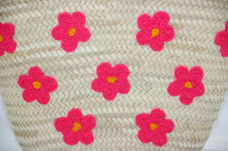 May include: Woven straw bag with bright pink flower appliqu&eacute;s. Each flower has a yellow center. The bag's weave is visible, creating a textured surface. The flowers are evenly spaced across the bag's surface.