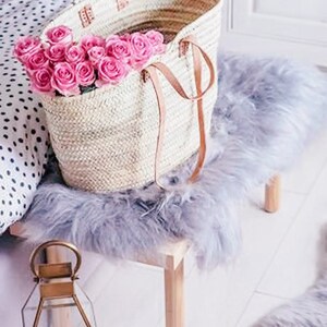 Puede incluir: Una bolsa de mano tejida de color blanco con correas de cuero llena de un ramo de rosas rosas. La bolsa est&aacute; sobre una superficie esponjosa de color gris claro.
