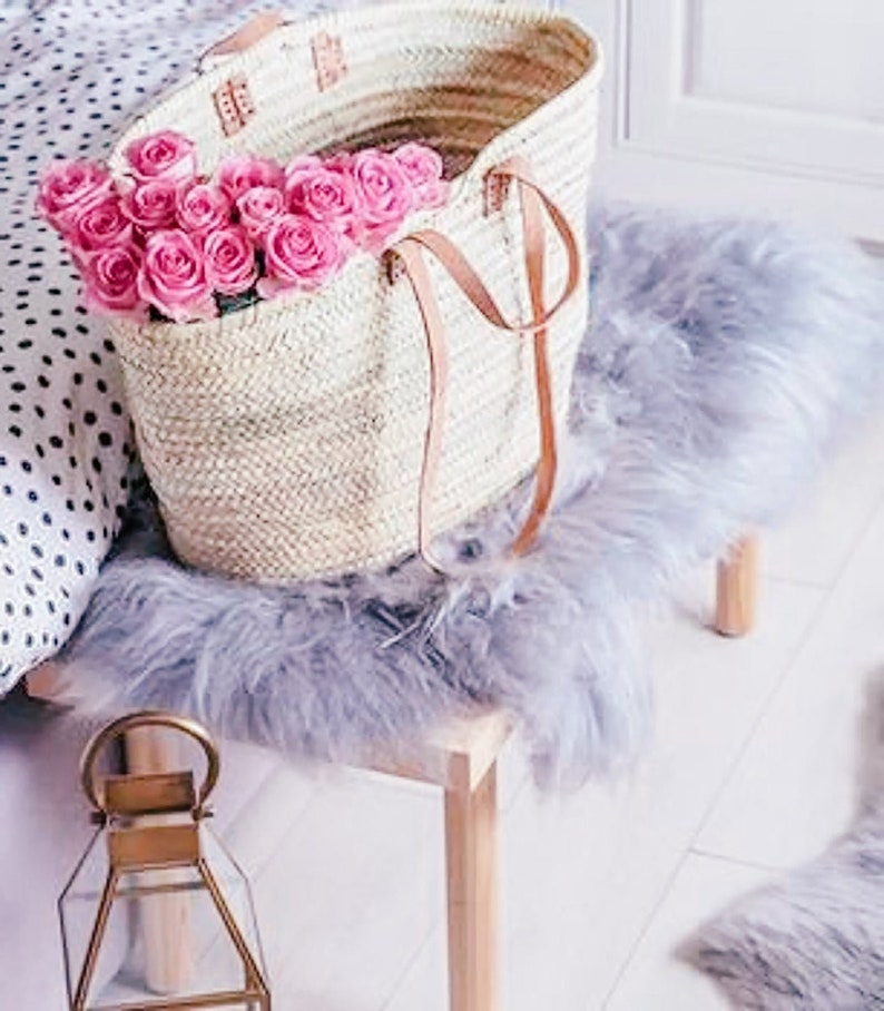 Puede incluir: Una bolsa de mano tejida de color blanco con correas de cuero llena de un ramo de rosas rosas. La bolsa est&aacute; sobre una superficie esponjosa de color gris claro.
