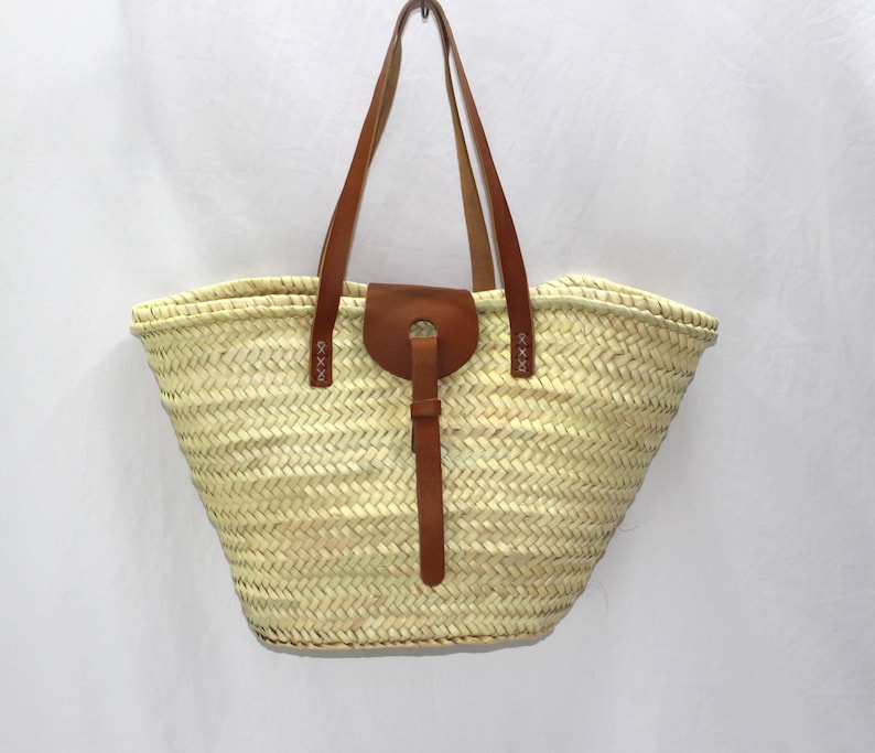 K&ouml;nnte beinhalten: Eine gro&szlig;e, hellbeige geflochtene Strohtasche mit braunen Ledergriffen und einem braunen Lederriemen mit Schnalle.