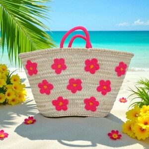 May include: A woven straw tote bag with bright pink flower appliqu&eacute;s and matching pink handles. The bag is on a sandy beach with yellow flowers and a turquoise ocean in the background.