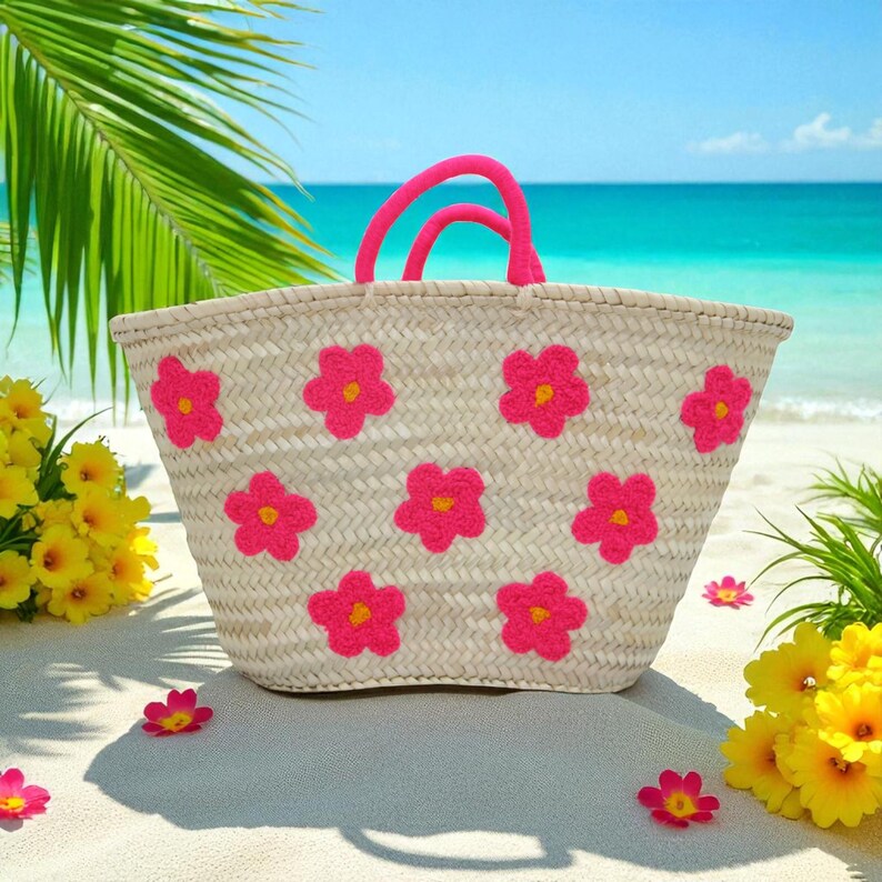 May include: A woven straw tote bag with bright pink flower appliqu&eacute;s and matching pink handles. The bag is on a sandy beach with yellow flowers and a turquoise ocean in the background.