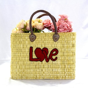 Op de afbeelding: Een strooien tas met bruine leren handvatten en een rood geborduurd "LOVE"-ontwerp. De tas is gevuld met roze en witte rozen.